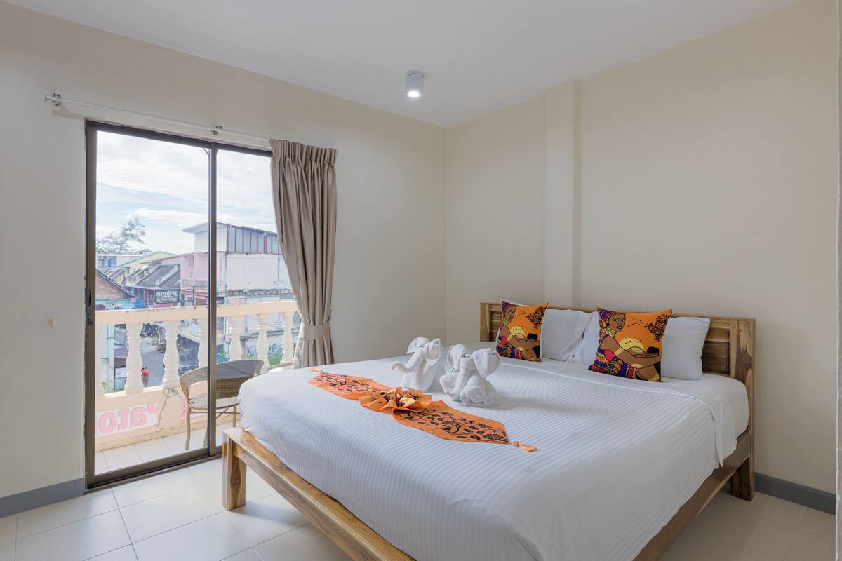 A hotel room with a large bed, a balcony with a view of buildings, and a wooden headboard.