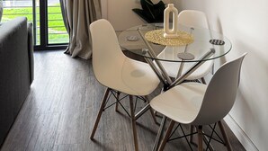 A modern dining area with a glass table, white chairs, and a view of greenery outside.