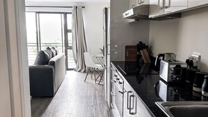 A modern kitchen with white cabinets, a stainless steel sink, and a coffee maker. There is a dining area with a white chair and a sofa.