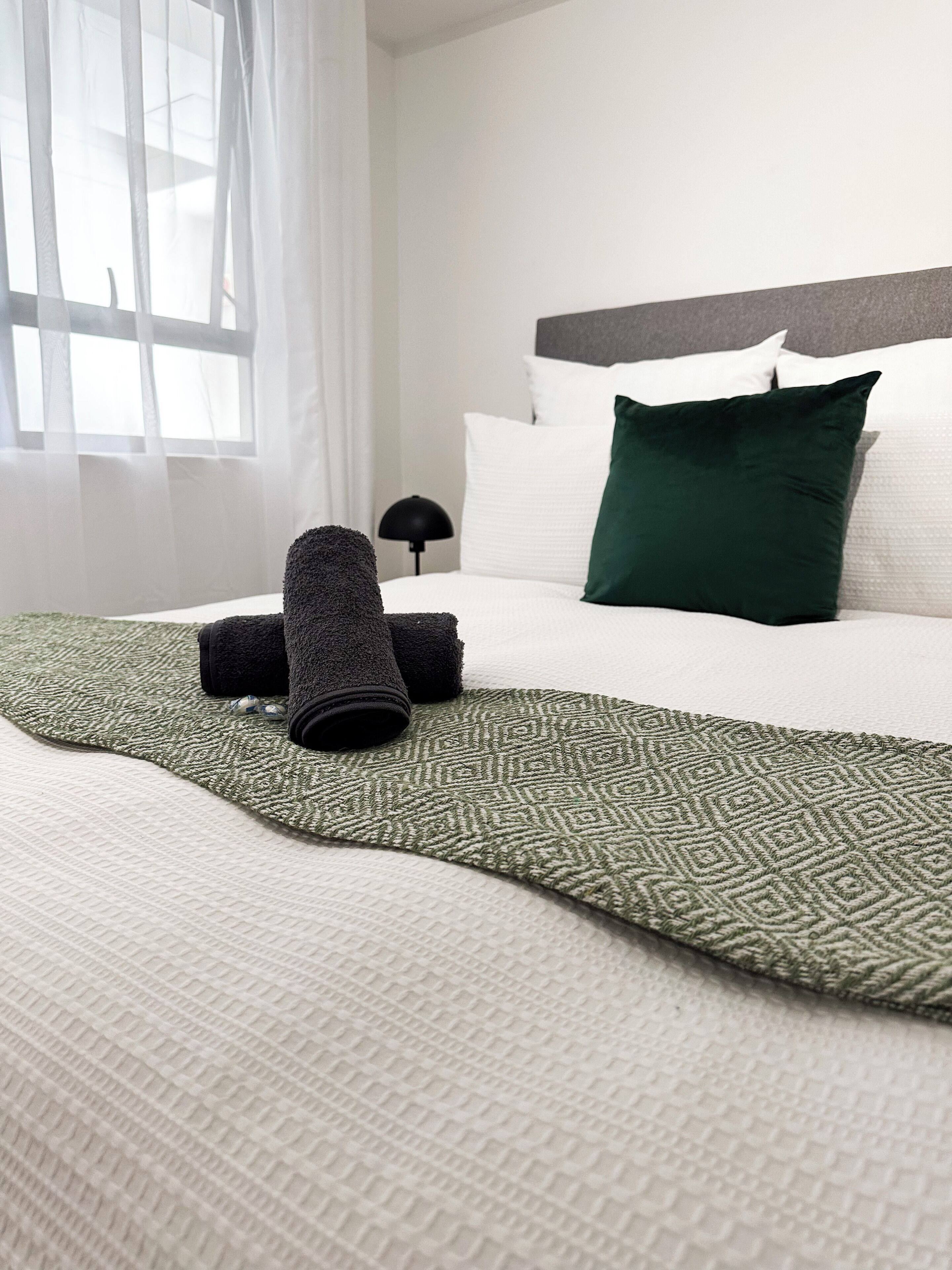 A neatly made bed with a green patterned bedspread, a dark green pillow, and rolled-up towels.