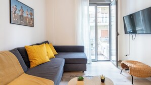 A modern living room with a grey sofa, yellow cushions, a wicker chair, a flat-screen TV, and a large window with a view of buildings. - The Beatles 2-BR Apartment in Athens (Athens)