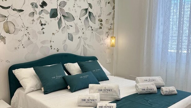 Ein ordentlich bezogenes Bett mit türkisfarbenen und weißen Bettwäsche, eine Wandmalerei mit floralem Muster und ein Fenster mit durchscheinenden Vorhängen.