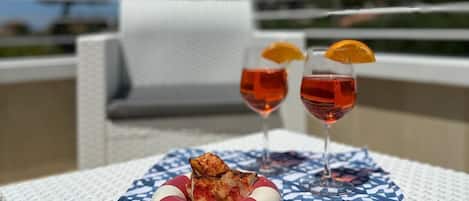 A wicker table with a patterned tablecloth, two glasses of red drink with orange slices, and a small dish with food on a balcony.