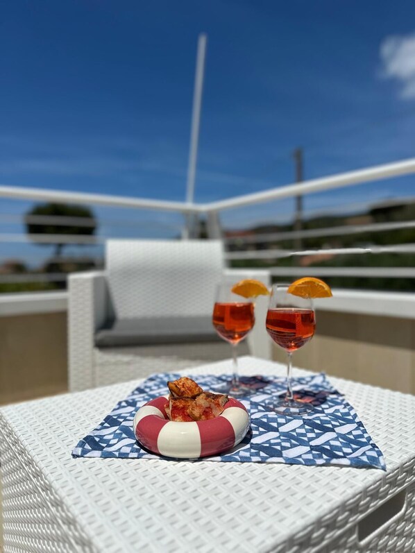 A wicker table with a patterned tablecloth, two glasses of red drink with orange slices, and a small dish with food on a balcony.