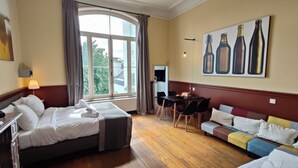Une chambre d’hôtel avec un lit, un bureau, un canapé, un tableau représentant des bouteilles et une fenêtre avec des rideaux.