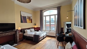 Quarto de hotel com cama, televisão, escrivaninha e janela grande.
