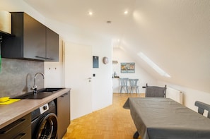 A compact room with a kitchenette, a washing machine, a dining area with chairs, and a bed.