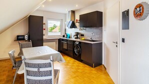 A compact kitchen with a microwave, oven, and washing machine, a dining area with a table and chairs, and a window providing natural light.
