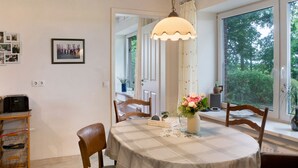 A dining area with a round table, chairs, a pendant light, a vase with flowers, and a window with a view of trees. - Country House 'Friesenhof' with Shared Terrace, Private Garden, and Wi-Fi (Wangerland)