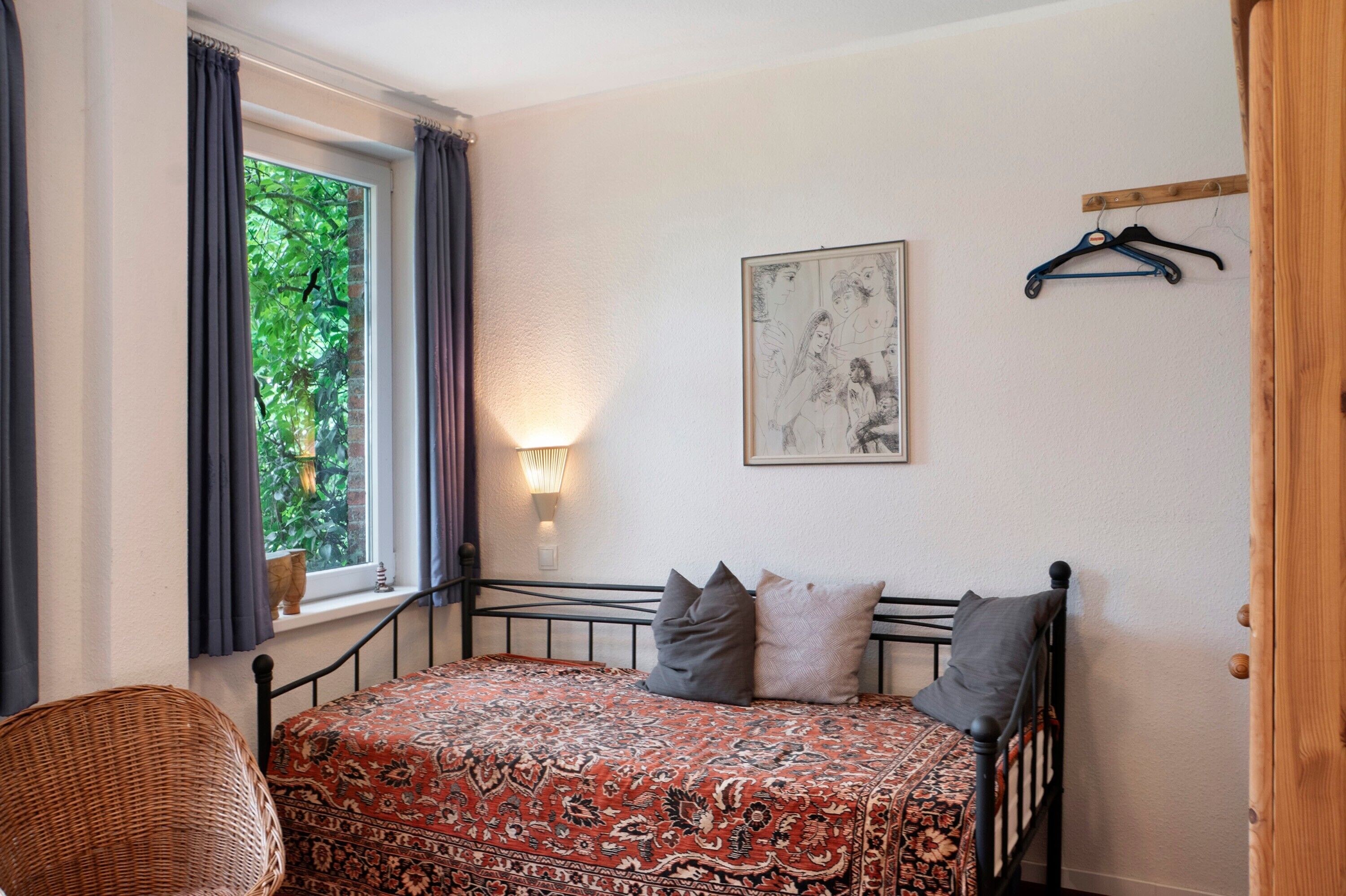 A bedroom with a bed, a window with curtains, a wall-mounted light, a hanging artwork, and a wooden door.