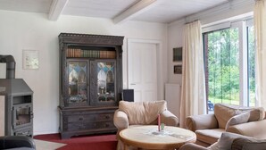 A living room with a fireplace, a large cabinet, a sofa, a round table, and a view of greenery through the window. - Country House 'Friesenhof' with Shared Terrace, Private Garden, and Wi-Fi (Wangerland)