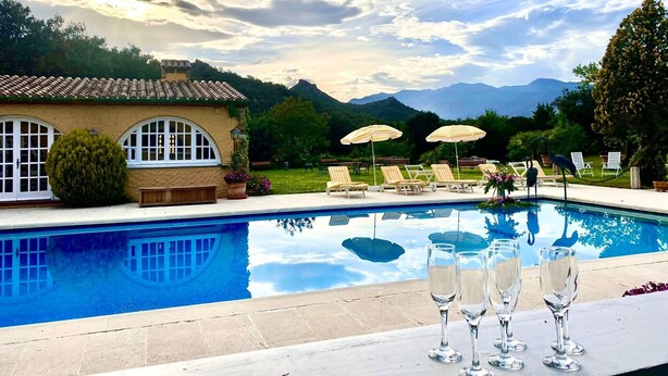 A pool with lounge chairs and umbrellas, a building with arched windows, and a mountainous backdrop.