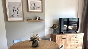 Un comedor con una mesa redonda de madera, seis sillas y un jarrón con flores. Hay dos obras de arte enmarcadas en la pared, un televisor sobre un mueble de madera, y suelo de madera.