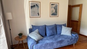 Una sala de estar con un sofá azul, dos cuadros enmarcados en la pared, una pequeña mesa redonda con una planta y suelo de madera.