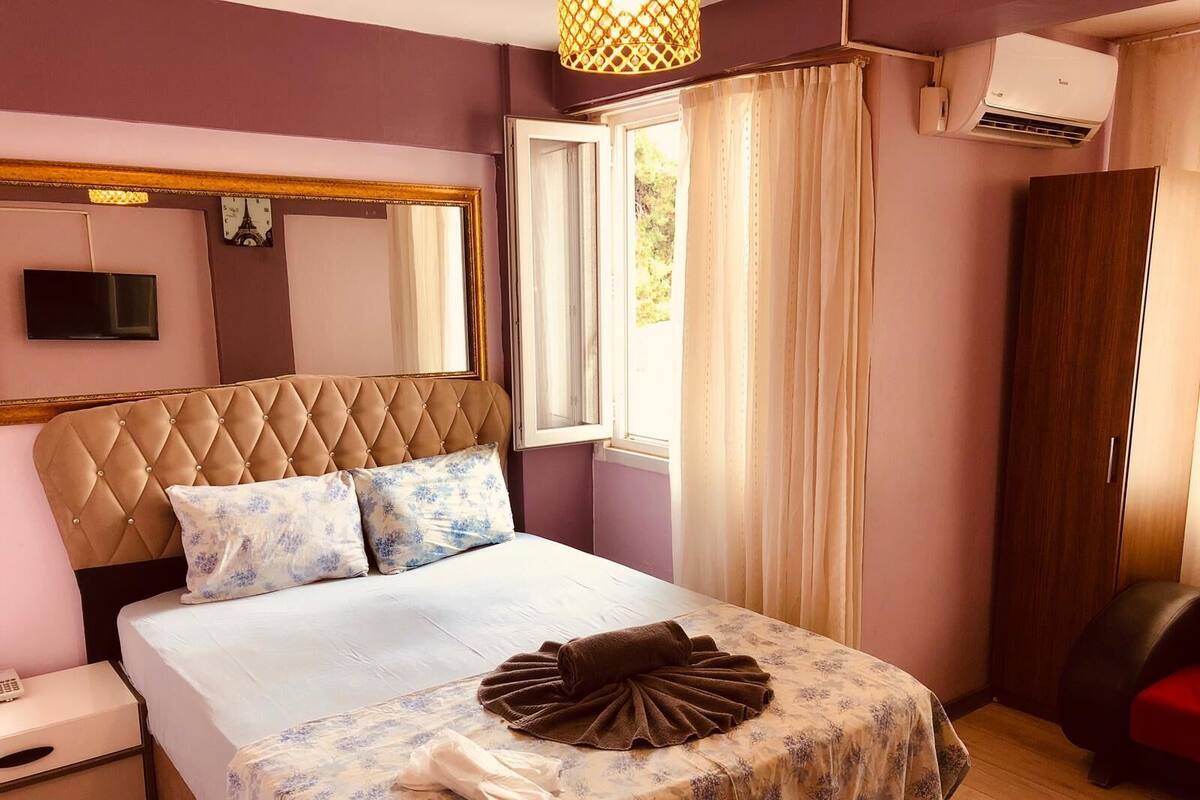 Ein Schlafzimmer mit einem gesteppten Kopfteil, einem Bett mit floraler Bettwäsche, einem Nachttisch mit Telefon, einem Fenster mit Vorhängen und einer Klimaanlage.
