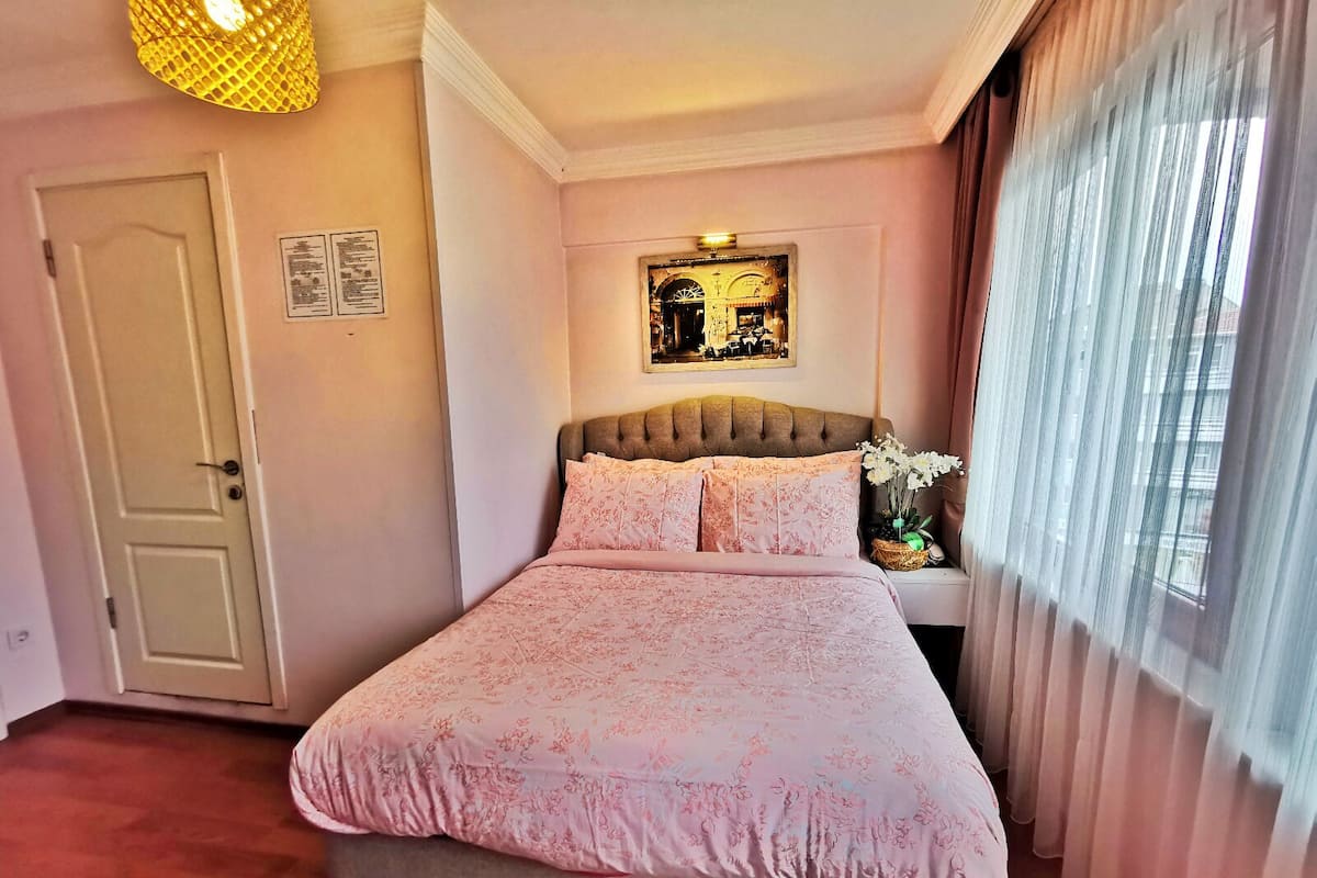 Ein Schlafzimmer mit Bett, Kopfstütze, Wandbild, Tür und Fenster mit Vorhängen.