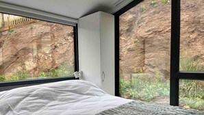 Una cama con ropa blanca, una manta estampada y vistas a un paisaje rocoso a través de amplias ventanas.