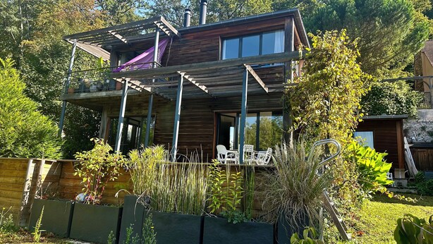 Une maison moderne à deux étages, avec une façade en bois, un patio couvert agrémenté de meubles d’extérieur et un jardin où poussent différentes espèces de plantes, le tout délimité par une clôture en bois.