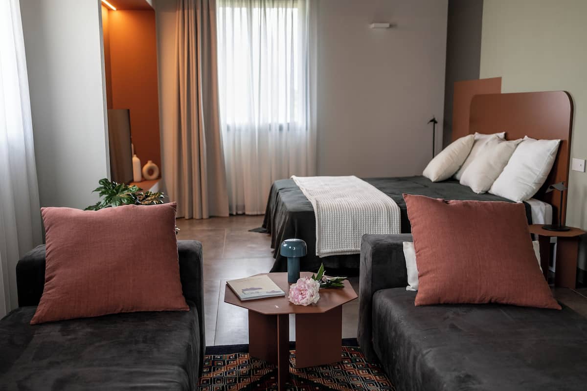 Ett modernt hotellrum med en säng, två fåtöljer, ett litet bord med en bok och en blomma, och ett fönster med gardiner.
