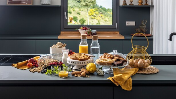 Un buffet per la colazione con diversi cibi, una finestra con vista sul verde e uno scaffale con oggetti decorativi.
