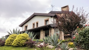 Una casa a due piani con facciata in pietra, un portico coperto e un giardino ricco di piante e fiori.