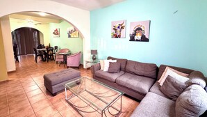 Una sala de estar con un sofá, un reposapiés y una mesa de centro de vidrio. Se observa un comedor con sillas y una mesa al fondo.