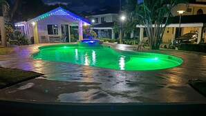 Vista nocturna de una piscina con iluminación verde, rodeada de palmeras y un edificio con zona cubierta.