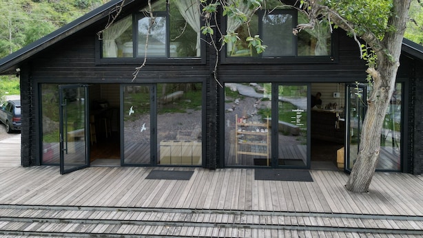 Eine moderne Holzhütte mit großen Glasüren und -fenstern, einer Holzterrasse und Blick auf einen Hügelhang.