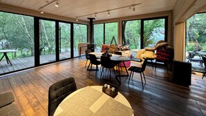 Ein geräumiges Zimmer mit Holzboden, großen Fenstern, einem Essbereich mit Tisch und Stühlen sowie einer gemütlichen Sitzecke mit Kissen.