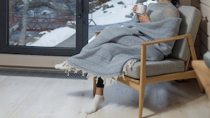 Eine Person sitzt an einem großen Fenster mit Vorhängen, eine Tasse haltend, in einem Zimmer mit Holzboden und einem schlichten Stuhl.