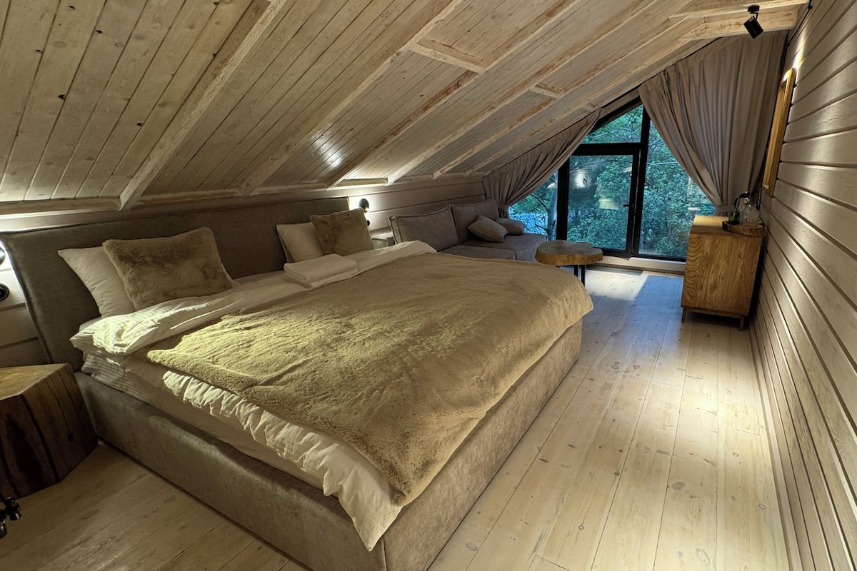 A cozy attic bedroom with a large bed, wooden walls, and a window overlooking greenery.