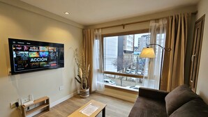 Una sala de estar con una televisión de pantalla plana montada en la pared, una mesa de centro de madera, un sofá marrón, un pequeño banco de madera y una planta en maceta.