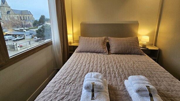 Habitación de hotel con una cama grande, dos almohadas, una mesita de noche con lámpara y vista a una iglesia por la ventana.