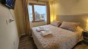 Habitación de hotel con cama, televisor montado en la pared, ventana con cortinas y mesita de noche con lámpara.