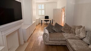 Ein Wohnzimmer mit einem großen Flachbildfernseher an der Wand, einem beigefarbenen Sofa mit Kissen, einem kleinen Tisch mit einem Sessel und einer Tür zu einem anderen Raum.