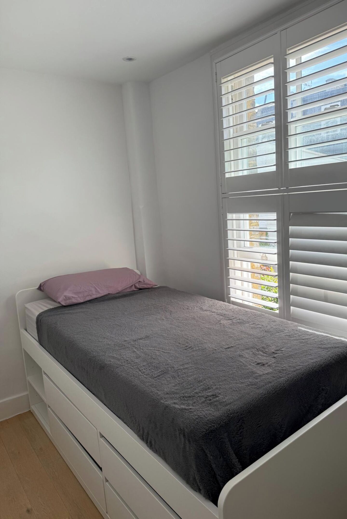 A small, single-bed room with a window and blinds, a white wall, and a wooden floor.