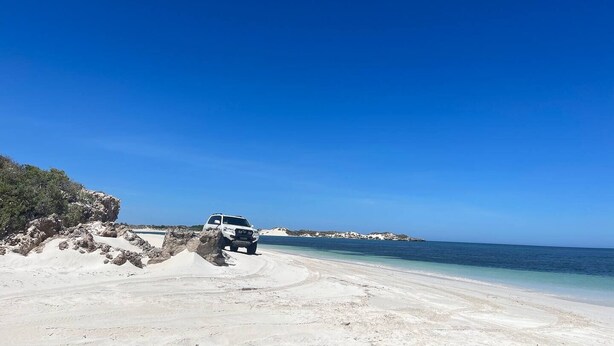 Un VUS blanc est garé sur une plage de sable, avec un ciel bleu dégagé et une mer calme en arrière-plan.