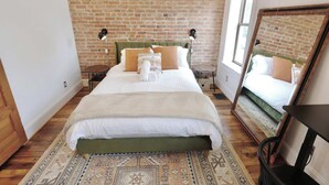 Een slaapkamer met een bakstenen muur, een bed met een groene hoofdeinde, twee nachtlampjes en een grote spiegel.