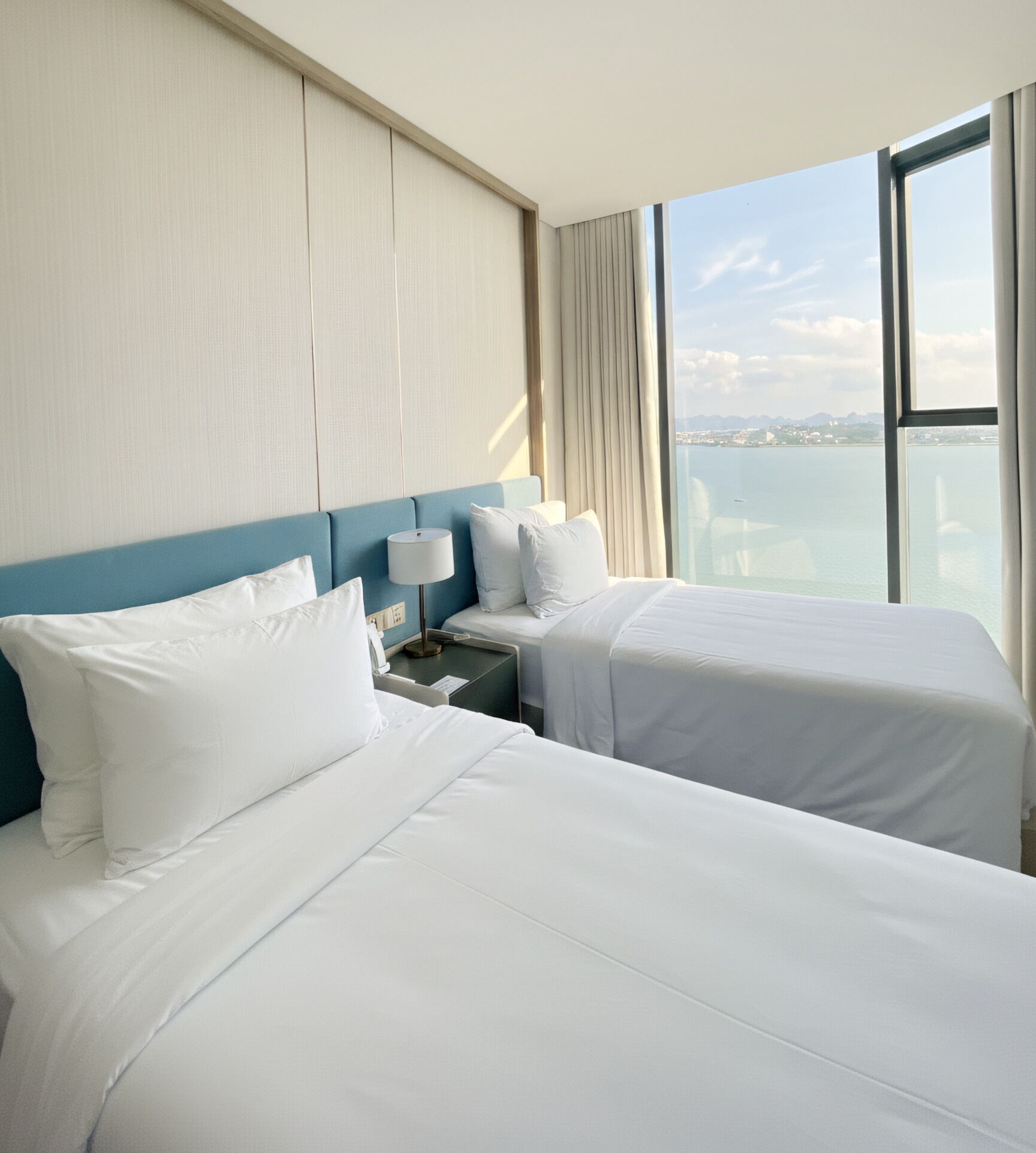 Ein Hotelzimmer mit zwei Betten, einem Nachttisch, einer Lampe und einem großen Fenster mit Blick auf das Meer.