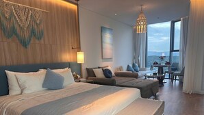 A modern hotel room with a large bed, a smaller bed, a sofa, a dining table, and a view of the mountains. - Alacarte Ha Long - Ha Apartment (Bai Chay)