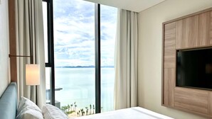 A hotel room with a large bed, a TV mounted on the wall, and a view of the sea through large windows. - Alacarte Ha Long - Ha Apartment (Bai Chay)