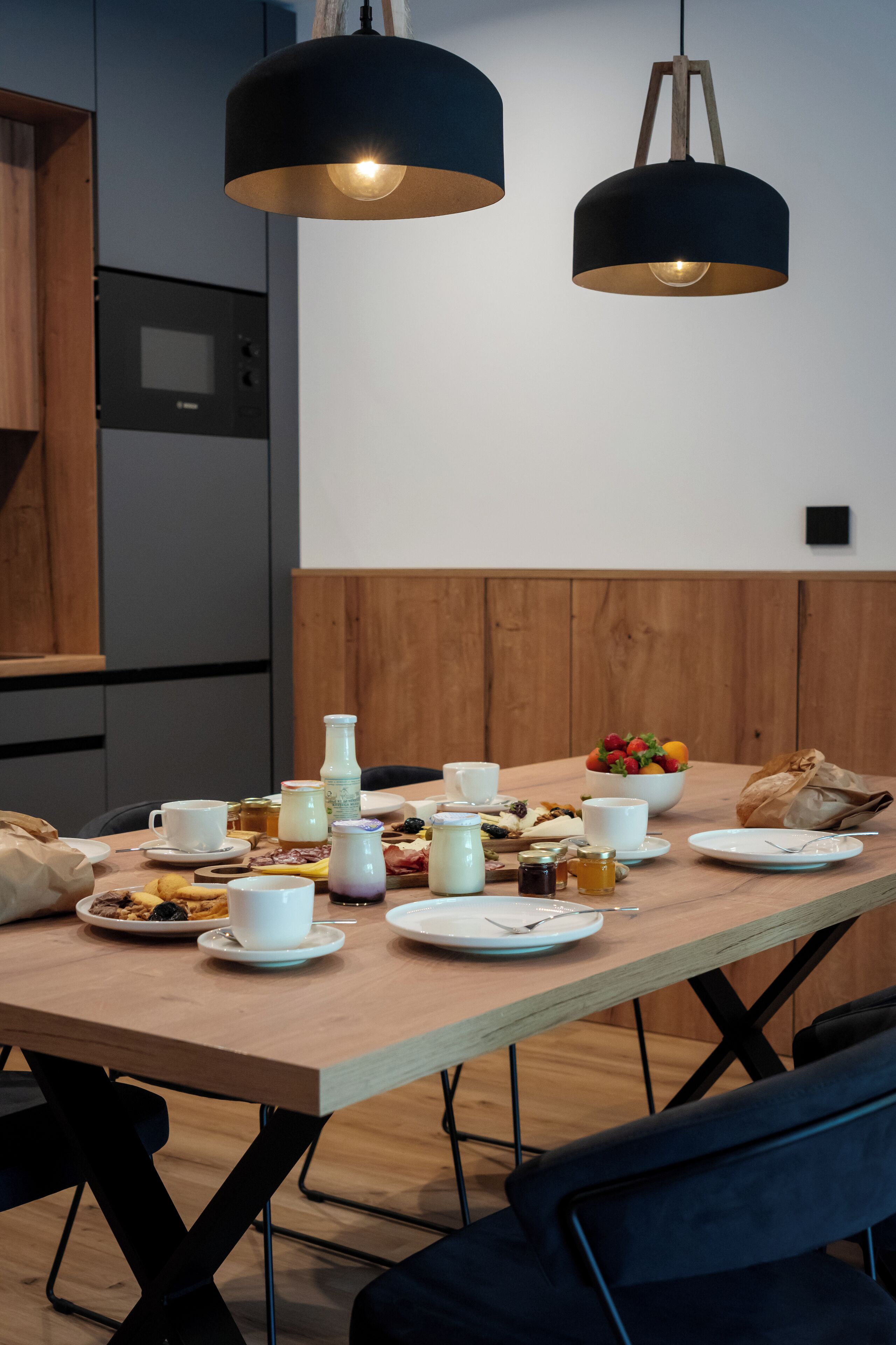 Una mesa de comedor dispuesta con alimentos para el desayuno, dos lámparas colgantes y un banco de madera.