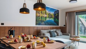 Ein modernes Wohnzimmer mit Essbereich, ausgestattet mit einem Holztisch, der für das Frühstück gedeckt ist, einer Couch und einem großen Fenster mit Blick auf eine Berglandschaft.