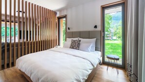 Ein modernes Schlafzimmer mit einem großen Bett, Blick auf Grünfläche und einem kleinen Tisch am Fenster.