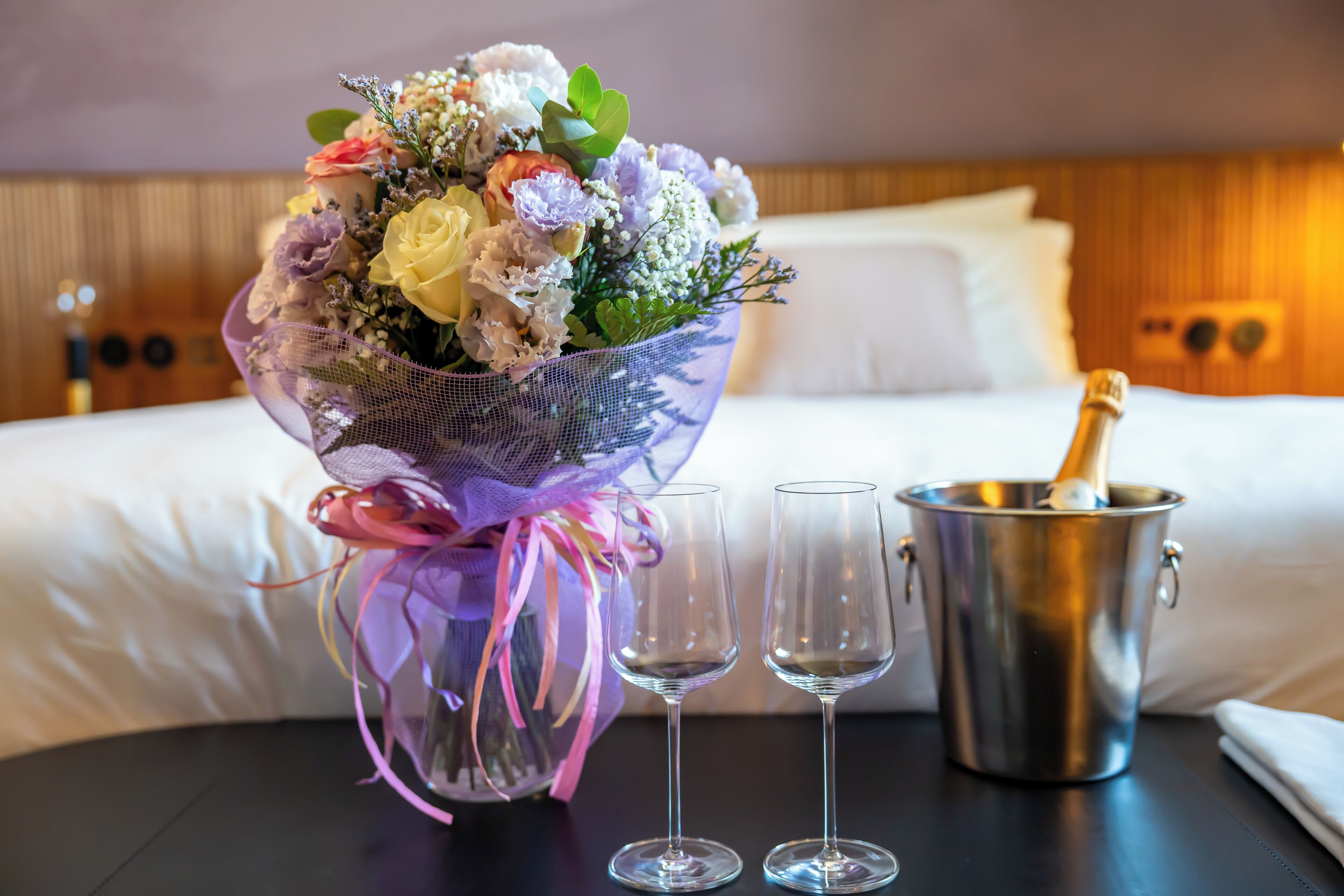 A bouquet of flowers in a decorative basket, two empty wine glasses, and a champagne bottle in a metal bucket on a table in a hotel room.