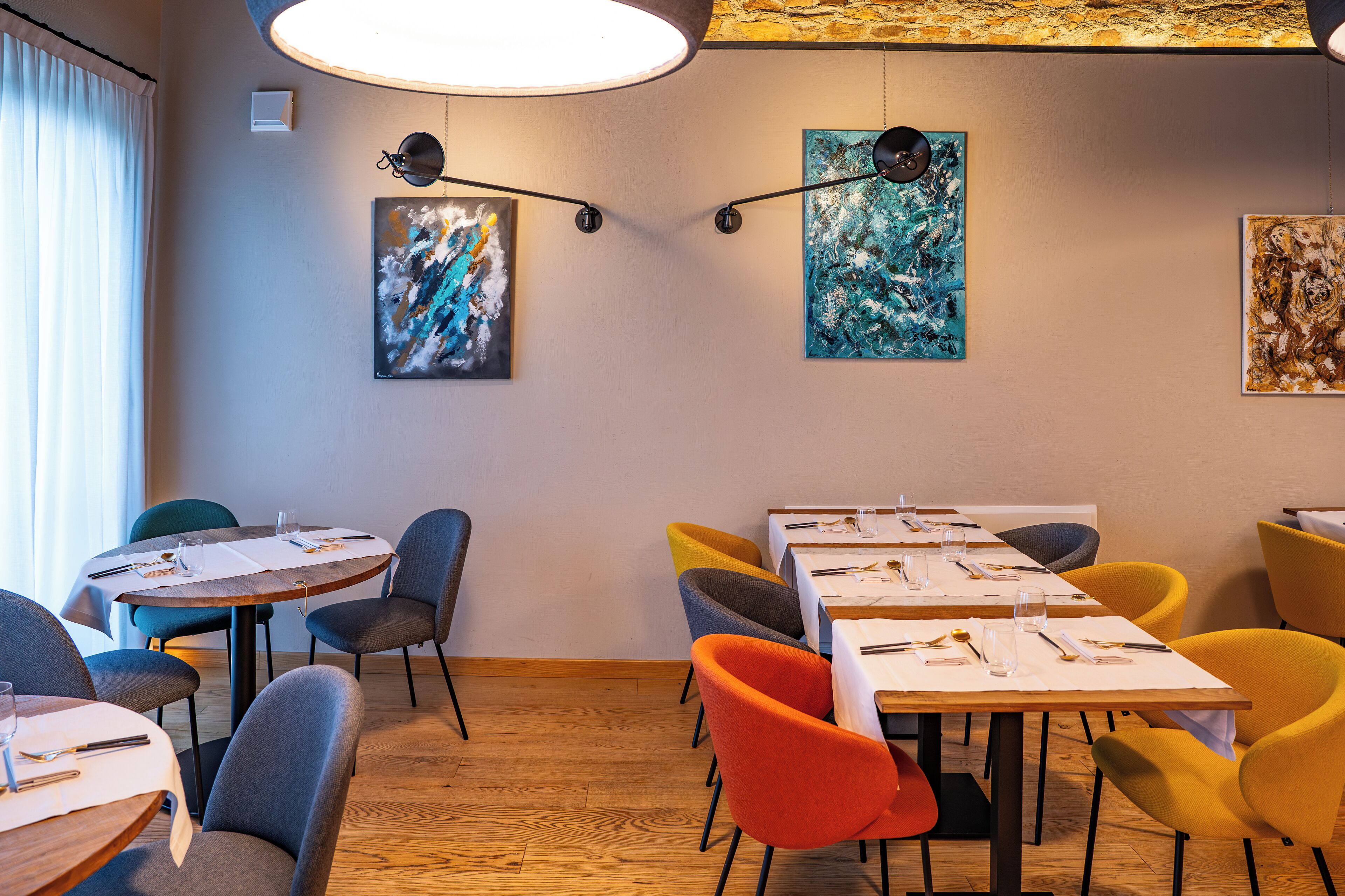 Un comedor con mesas redondas y rectangulares, sillas de varios colores y pinturas abstractas en las paredes.