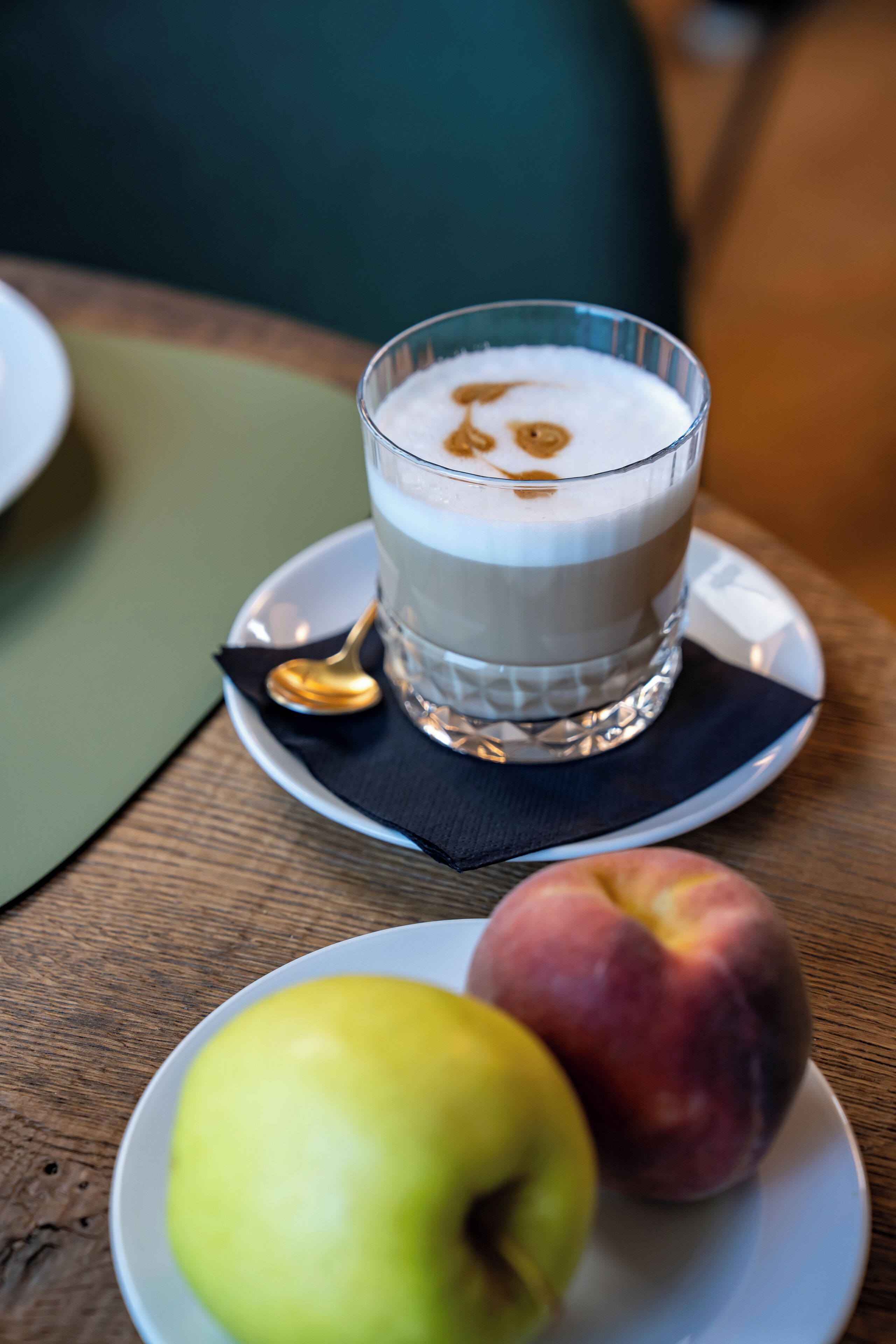 Ein Glas mit einem geschichteten Getränk auf einer Untertasse, zusammen mit einem Löffel und einem Teller mit einem Apfel und einem Pfirsich.