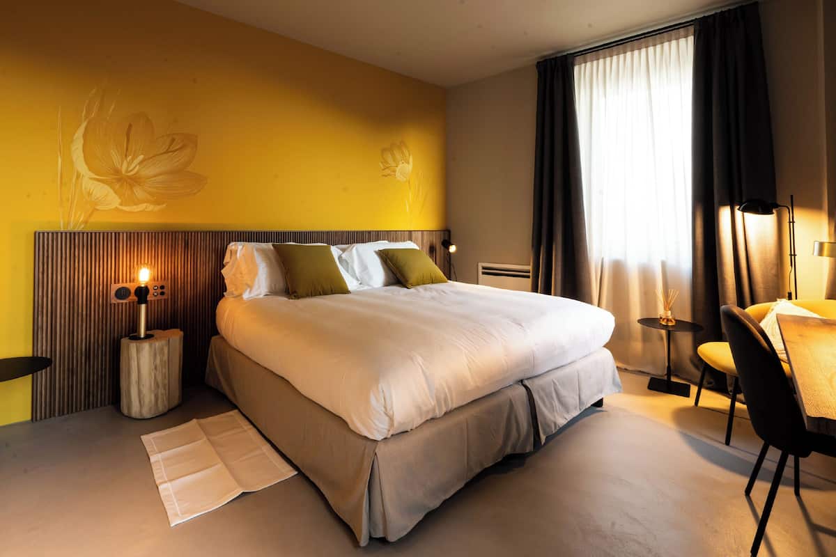 A hotel room with a large bed, a bedside table with a lamp, a chair, and a desk with a lamp.