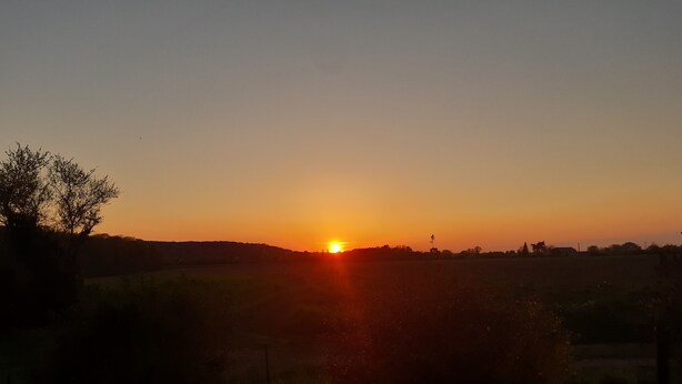 Een zonsondergang boven een landelijk gebied met velden en enkele bomen.