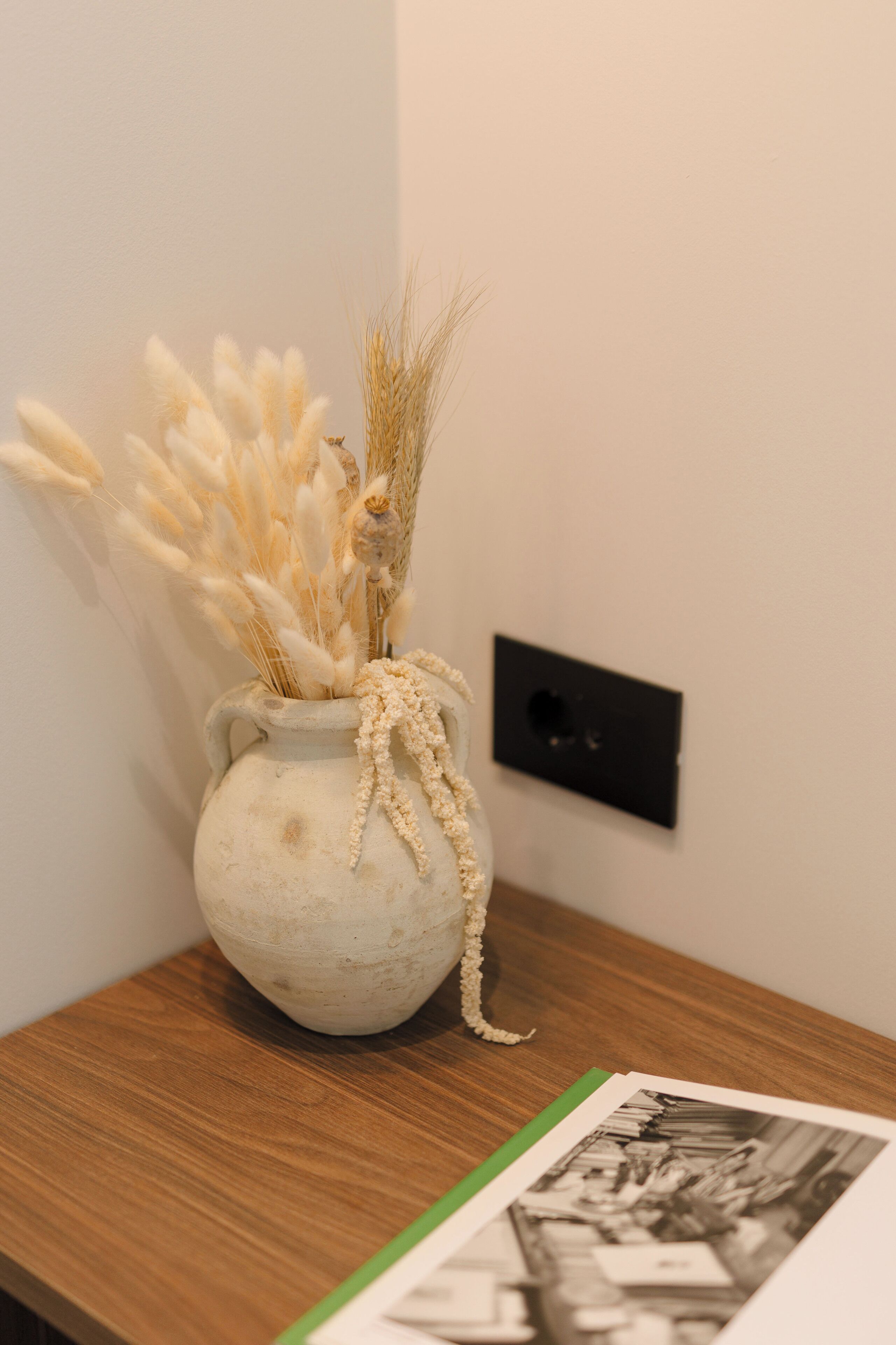 Eine Vase mit getrockneten Pflanzen auf einem Holztisch vor weißer Wand.